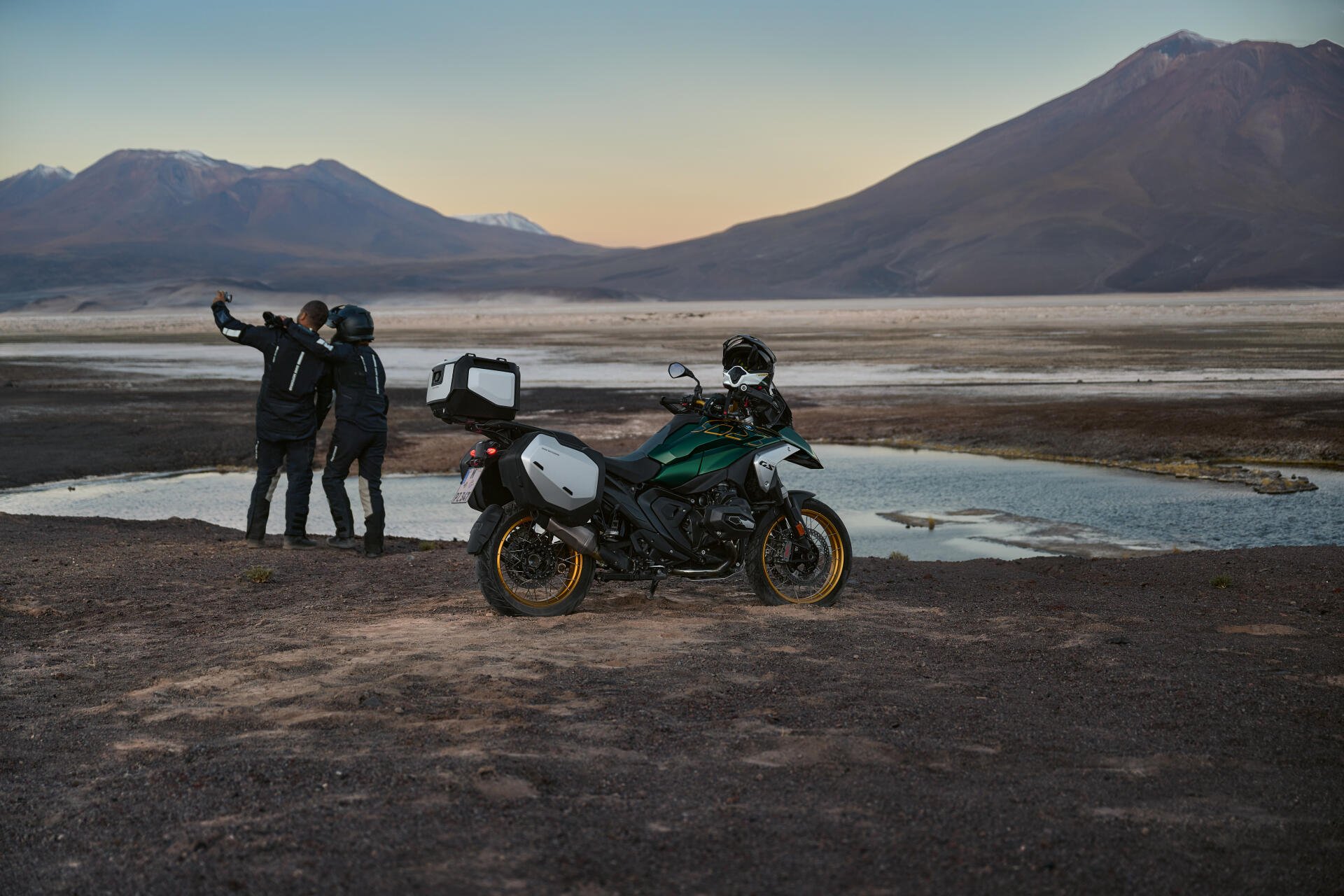 A BMW R1300GS motorcycle parked near a tranquil water body, with two riders capturing a scenic sunset backdrop of mountains. 4K Ultra HD wallpaper showcasing adventure.