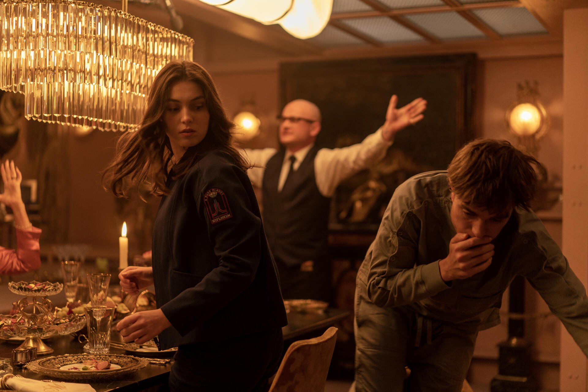 A scene from the 2025 movie Mickey 17, featuring Robert Pattinson and Anamaria Vartolomei. The image captures a tense moment at a dinner table, illuminated by warm lighting.