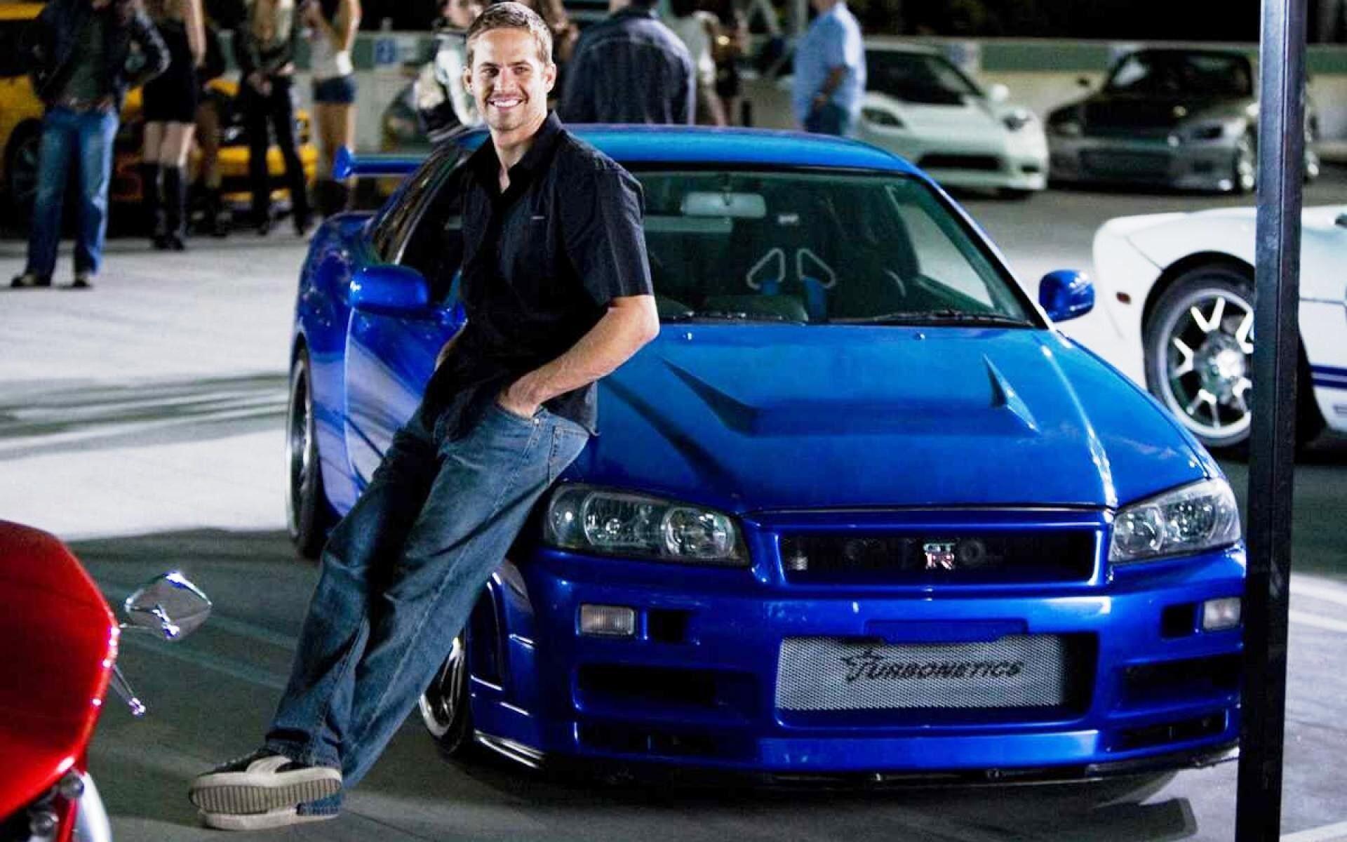 Brian O'Conner leaning against a blue Nissan Skyline R34, featured in Veloces e Furiosos 4, captured as an HD PC desktop wallpaper and background.