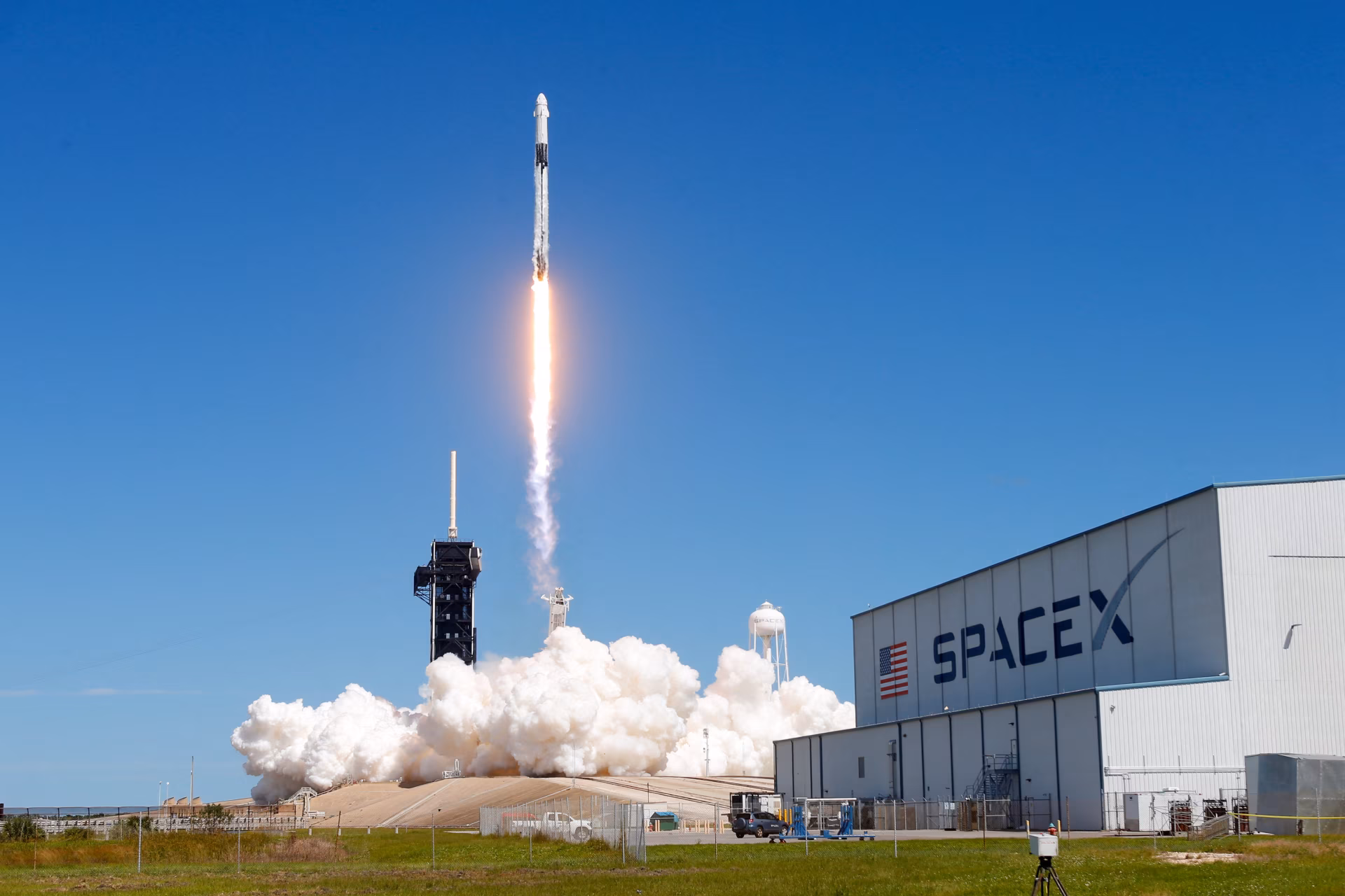 A SpaceX rocket launches against a clear blue sky, with billowing clouds of smoke at the launch site, showcasing man-made advancement in space exploration. 4K Ultra HD desktop wallpaper.