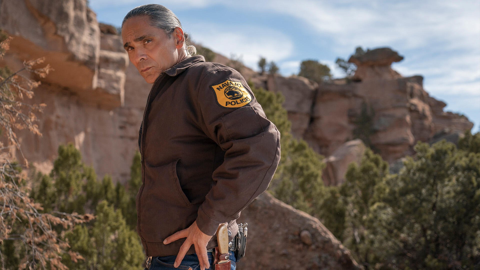 Zahn McClarnon in character as a law enforcement officer in Dark Winds, set against a striking desert landscape, showcasing the show's captivating atmosphere. HD wallpaper.