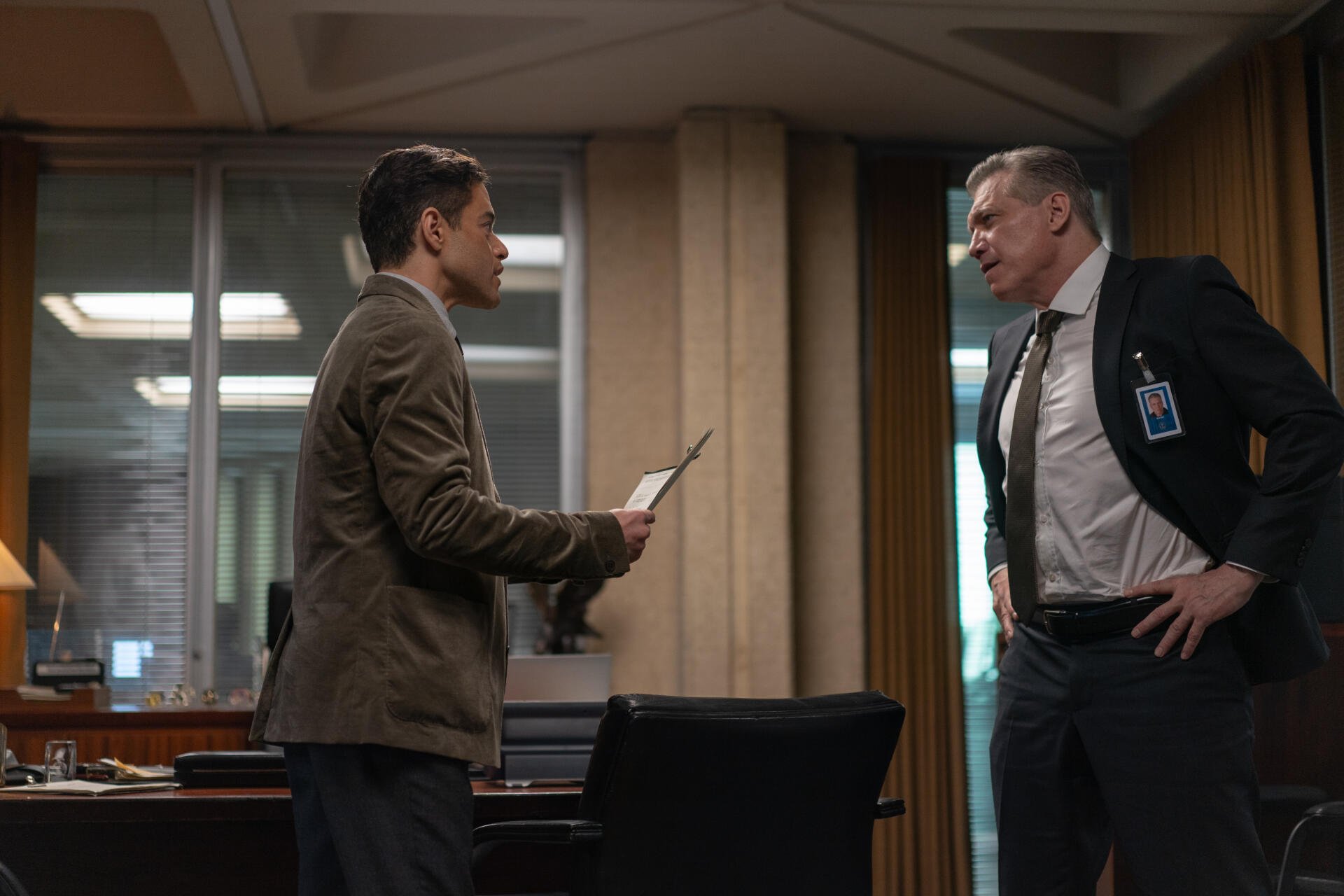 Scene from the movie The Amateur (2025) featuring Rami Malek and Holt McCallany engaged in a tense discussion in an office setting. 4K Ultra HD wallpaper for desktop backgrounds.