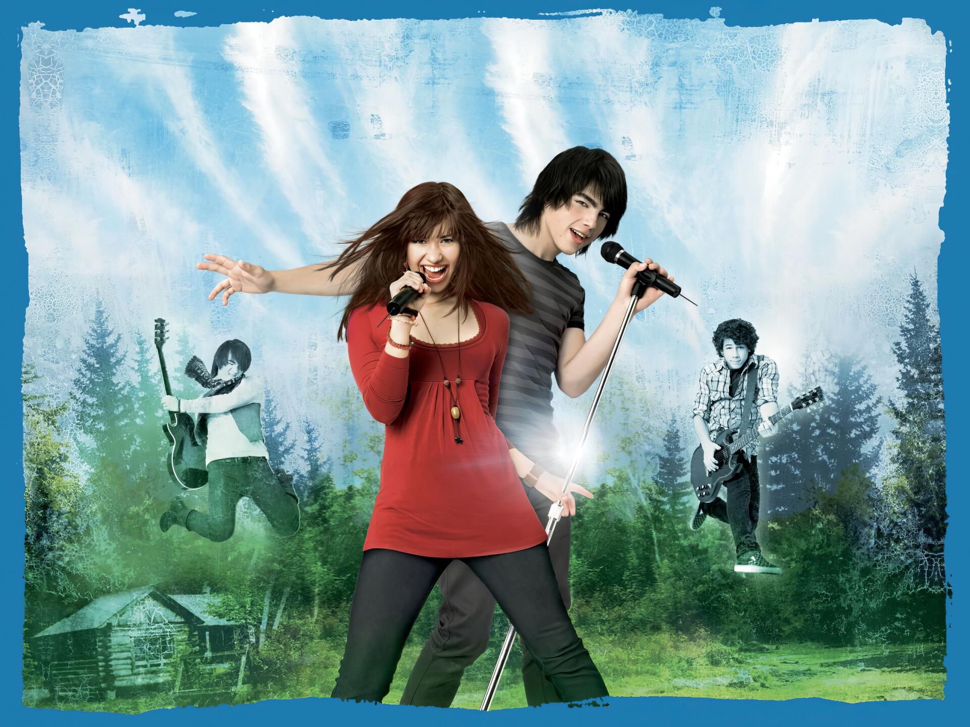 HD PC desktop wallpaper of Camp Rock: two young singers with microphones center stage, sunlight rays over a forest camp and band members playing in the misty background.