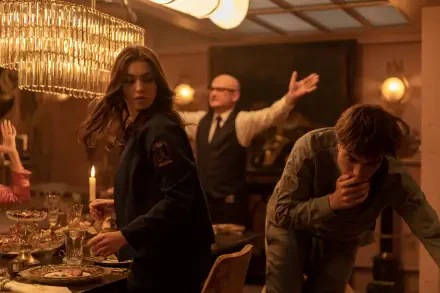 A scene from the 2025 movie Mickey 17, featuring Robert Pattinson and Anamaria Vartolomei. The image captures a tense moment at a dinner table, illuminated by warm lighting.