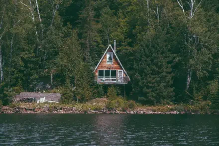 A wooden cottage nestled among dense forest trees on a calm coastal shore, captured in a high-definition desktop wallpaper and background.
