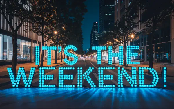 A vibrant 4K Ultra HD wallpaper featuring the text IT'S THE WEEKEND! illuminated in blue, set against a lively city backdrop at night.