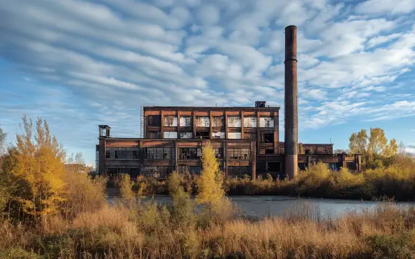 A striking abandoned factory stands by a calm body of water, surrounded by golden foliage and under a dramatic sky, making a captivating 4K Ultra HD desktop wallpaper.