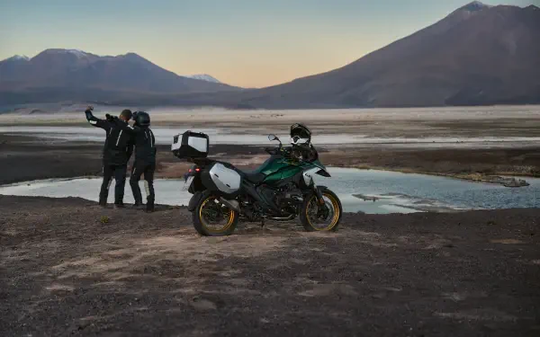 A BMW R1300GS motorcycle parked near a tranquil water body, with two riders capturing a scenic sunset backdrop of mountains. 4K Ultra HD wallpaper showcasing adventure.