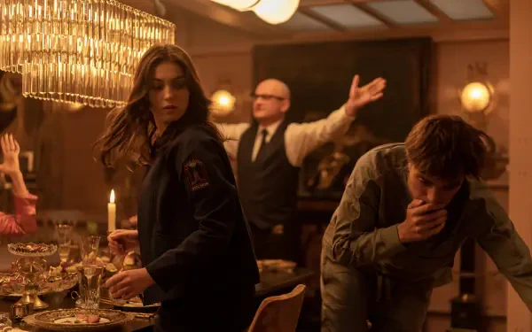 A scene from the 2025 movie Mickey 17, featuring Robert Pattinson and Anamaria Vartolomei. The image captures a tense moment at a dinner table, illuminated by warm lighting.