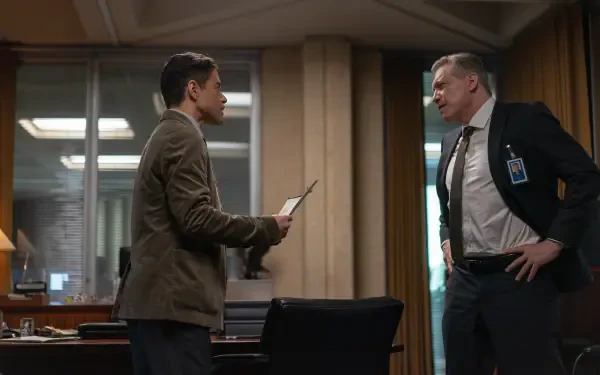 Scene from the movie The Amateur (2025) featuring Rami Malek and Holt McCallany engaged in a tense discussion in an office setting. 4K Ultra HD wallpaper for desktop backgrounds.
