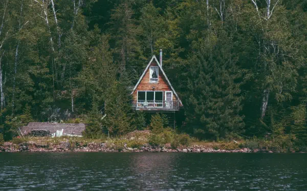 A wooden cottage nestled among dense forest trees on a calm coastal shore, captured in a high-definition desktop wallpaper and background.
