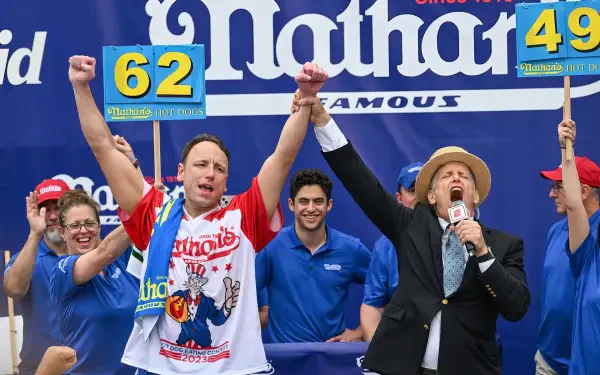 Joey Chestnut celebrates his victory at a Nathan's hot dog eating competition with raised arms and a crowd cheering behind him in this HD desktop wallpaper.