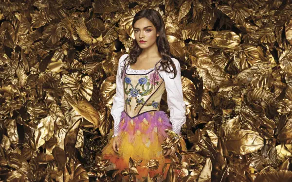 Woman in a colorful feathered costume amid a backdrop of golden leaves — The Hunger Games: The Ballad of Songbirds & Snakes 8K Ultra HD PC desktop wallpaper.