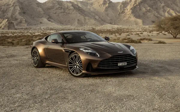 Brown Aston Martin DB12 parked on sunlit desert gravel with jagged mountains in the background — 4K Ultra HD PC desktop wallpaper.