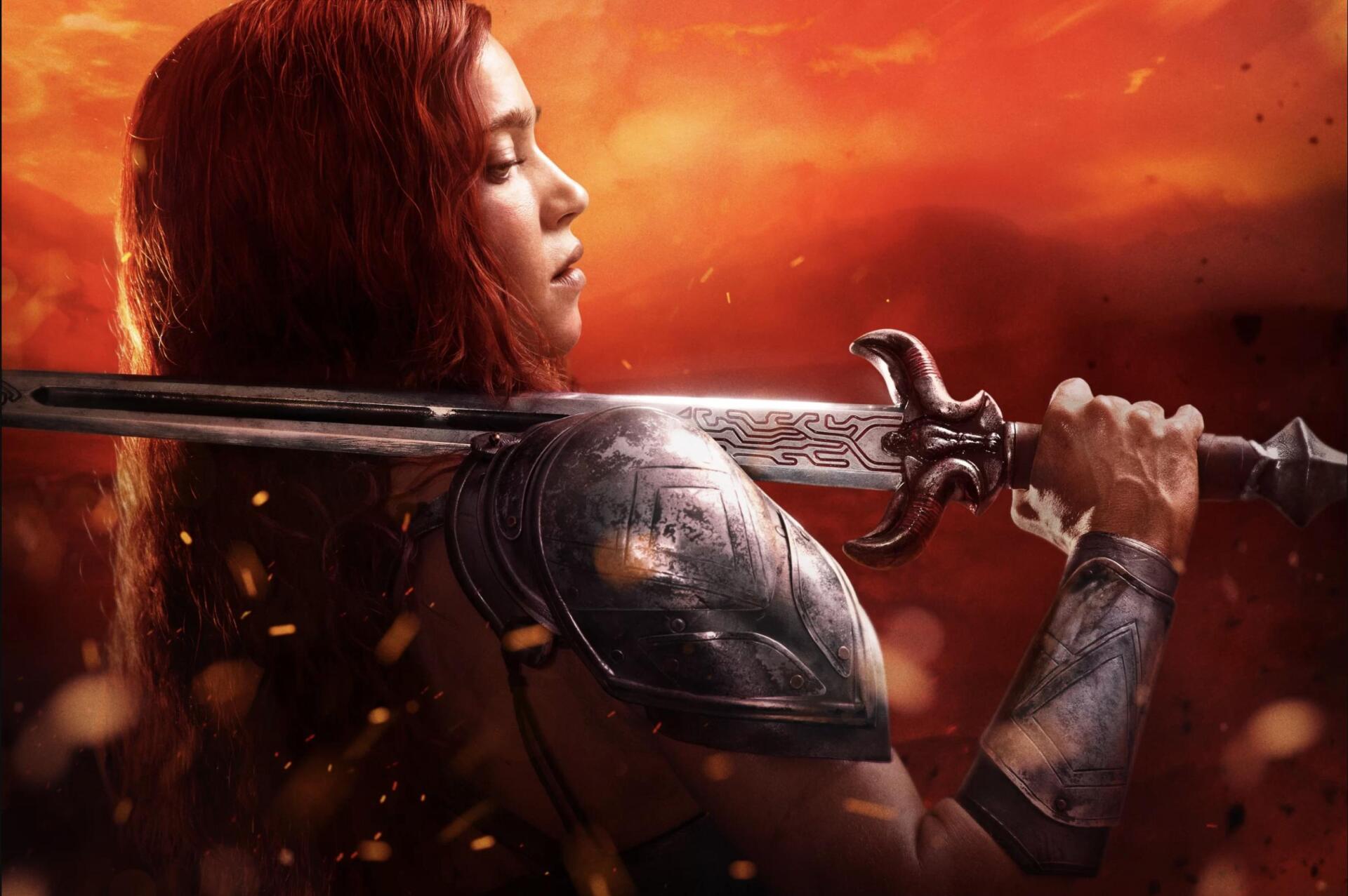 HD PC desktop wallpaper from the Red Sonja movie: red-haired woman warrior in armor gripping a broad sword against a fiery orange sky background.