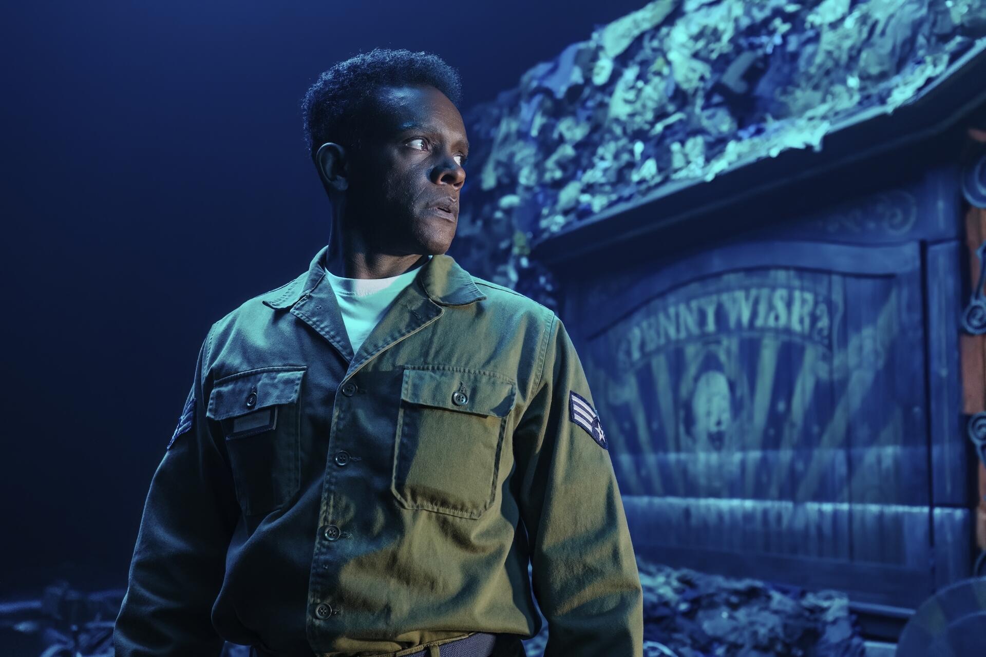 HD PC desktop wallpaper from the TV show It: Welcome to Derry — a man in a military jacket looks past a faded PENNYWISE poster under eerie moonlight.
