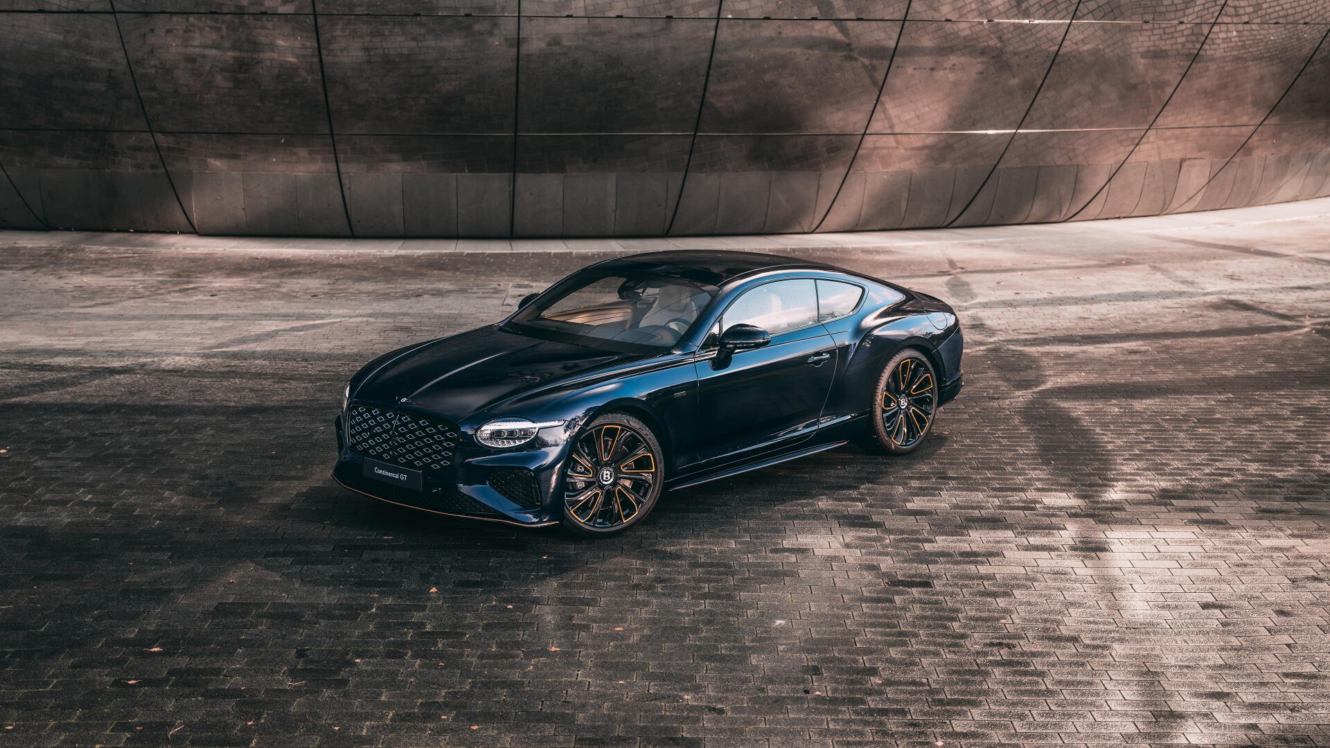 Sleek dark blue Bentley Continental GT luxury car on wet cobblestone in front of a curved concrete wall — 8K Ultra HD PC desktop wallpaper