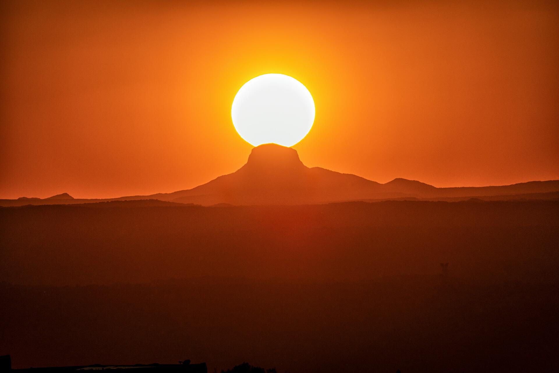 Silhouette of distant dunes and a central peak beneath a huge sun at desert sunset with an orange sky — 5K Ultra HD PC desktop wallpaper/background.