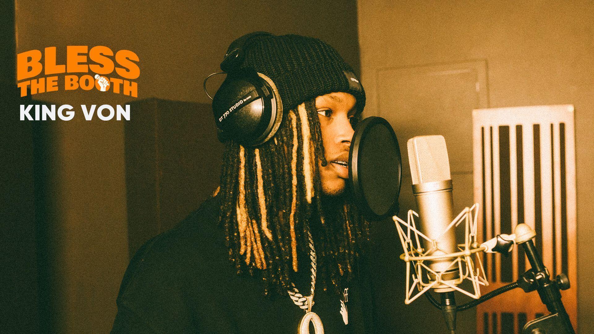 HD PC desktop wallpaper of a rapper in a studio booth—wearing a beanie and headphones, braided hair, singing into a large condenser mic with a warm, orange-lit background.
