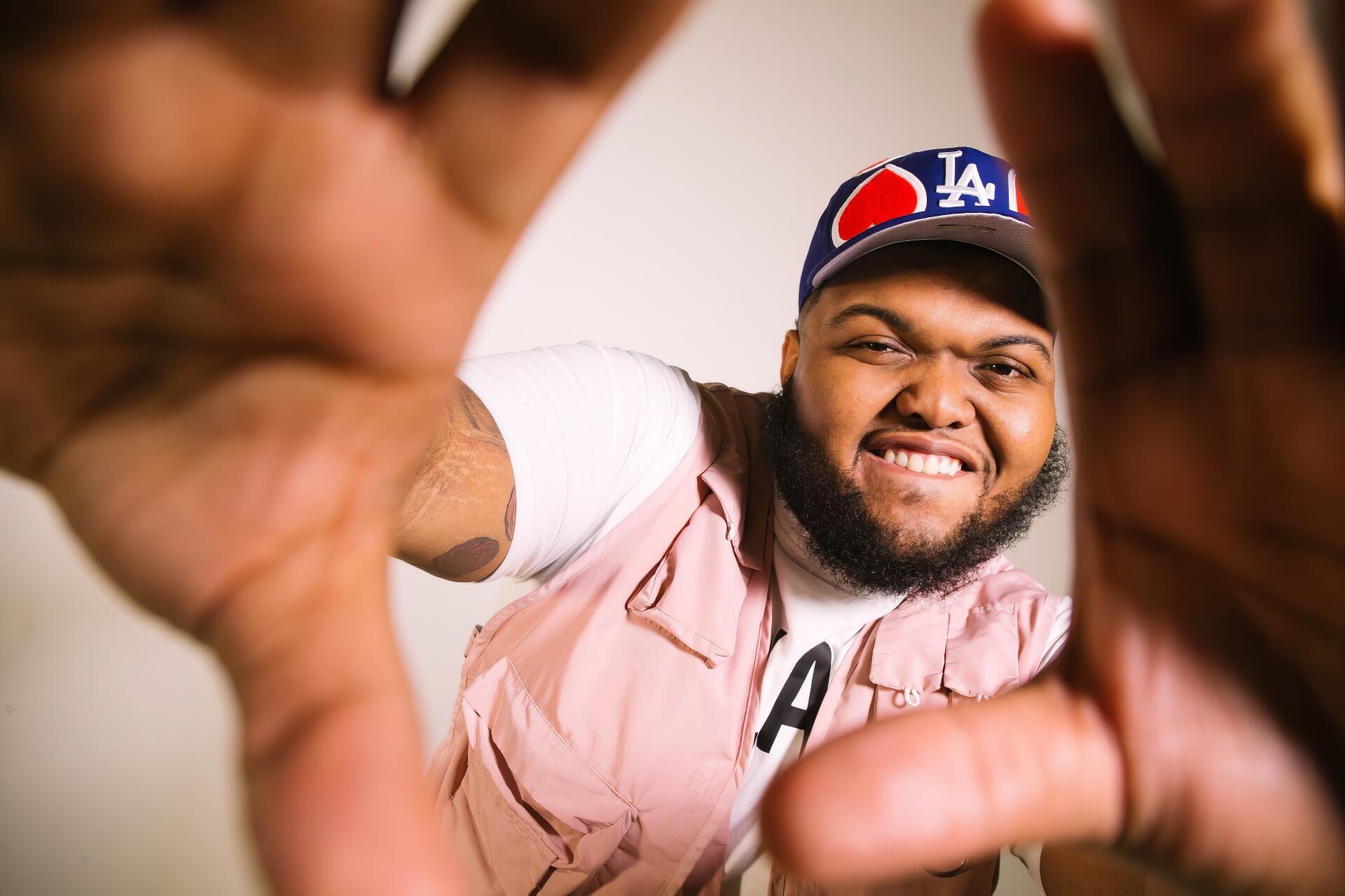 HD PC desktop wallpaper of an American YouTuber smiling and reaching toward the camera, hands framing the shot, wearing an LA cap and pink vest.