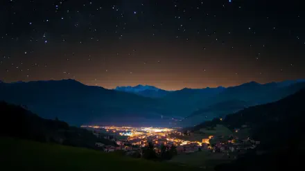 HD desktop wallpaper: serene mountain landscape at night, a starry sky over a glowing valley town nestled between dark peaks.