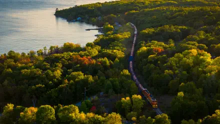 5K Ultra HD desktop wallpaper: aerial landscape view of a train on a winding railroad cutting through dense green forest beside a calm lake.