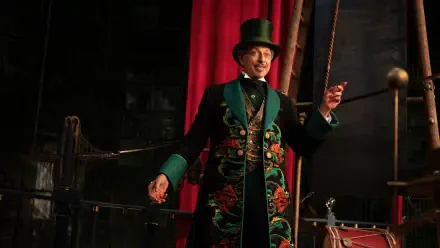 4K Ultra HD PC desktop wallpaper: an actor from the movie Wicked: For Good in a floral embroidered coat and top hat, smiling and gesturing on a dim, theatrical stage.