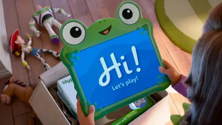Toy Story 5 5K Ultra HD PC Desktop Wallpaper: a child holds a frog-shaped tablet that says Hi! Let's play! with Woody, Buzz and other toys in a box on a wooden floor.