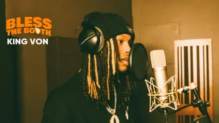 HD PC desktop wallpaper of a rapper in a studio booth—wearing a beanie and headphones, braided hair, singing into a large condenser mic with a warm, orange-lit background.