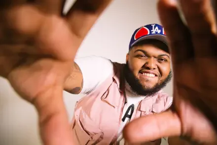 HD PC desktop wallpaper of an American YouTuber smiling and reaching toward the camera, hands framing the shot, wearing an LA cap and pink vest.