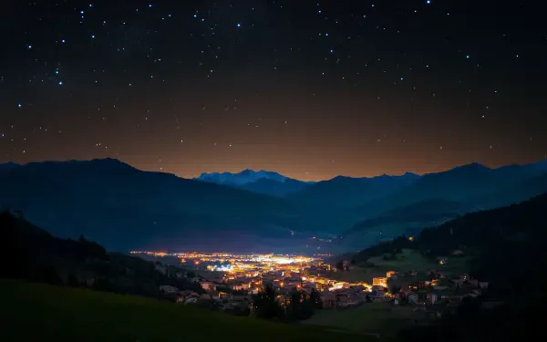 HD desktop wallpaper: serene mountain landscape at night, a starry sky over a glowing valley town nestled between dark peaks.