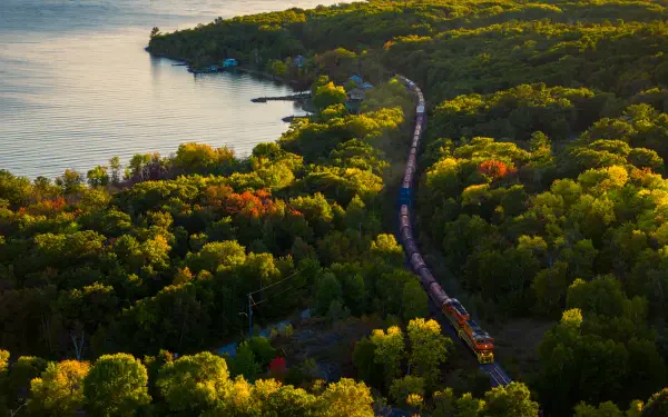 5K Ultra HD desktop wallpaper: aerial landscape view of a train on a winding railroad cutting through dense green forest beside a calm lake.