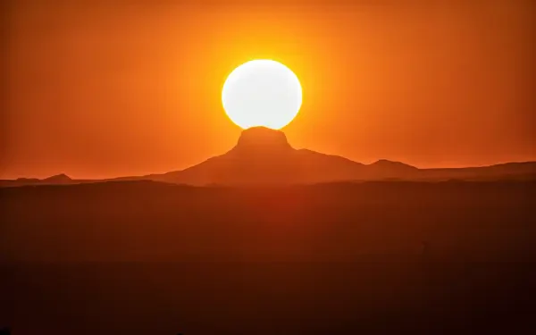 Silhouette of distant dunes and a central peak beneath a huge sun at desert sunset with an orange sky — 5K Ultra HD PC desktop wallpaper/background.