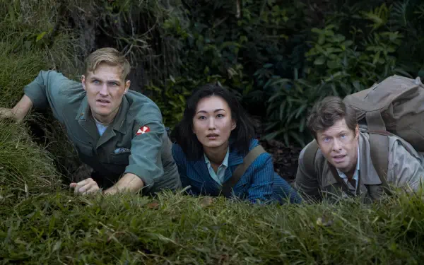 Three characters crouch in tall grass, tense and alert amid a dense forest — Monarch: Legacy of Monsters 5K Ultra HD PC desktop wallpaper and background.