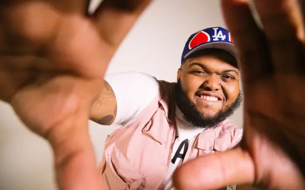 HD PC desktop wallpaper of an American YouTuber smiling and reaching toward the camera, hands framing the shot, wearing an LA cap and pink vest.