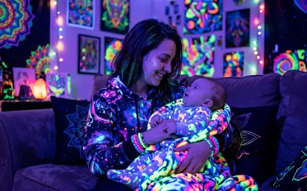 Mother and baby on a couch, mother cradling infant amid neon blacklight psychedelic art and fairy lights; vivid glowing colors. 5K Ultra HD PC desktop wallpaper and background.