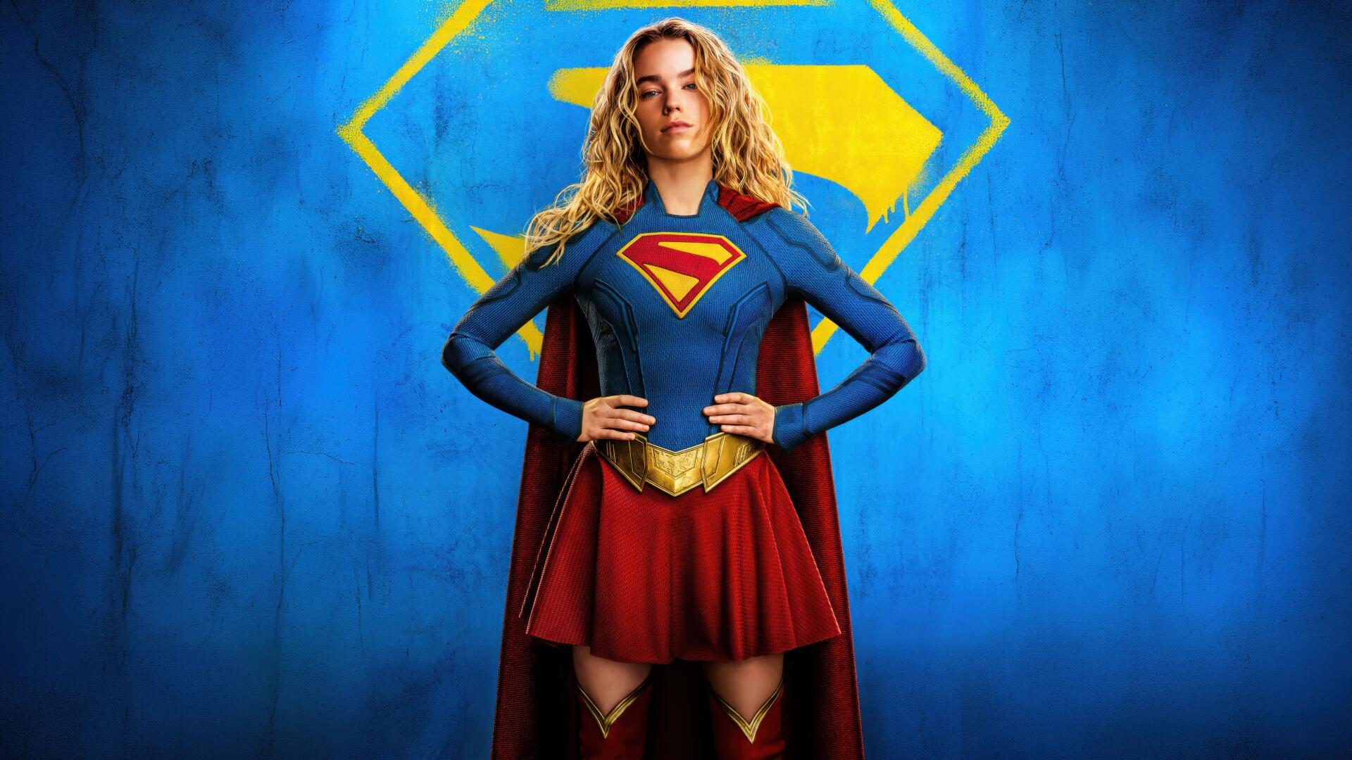 Supergirl (2026) 4K Ultra HD PC desktop wallpaper: Supergirl stands hands on hips in blue suit and red cape, emblem glowing behind her against a vivid blue background.