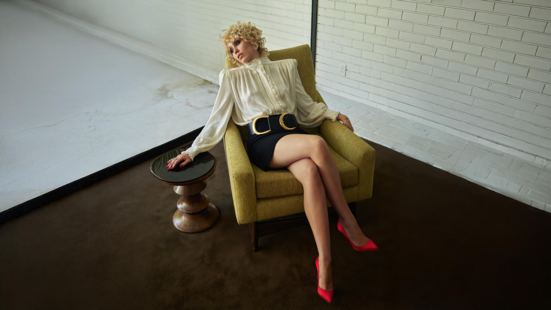 Blonde female pop singer and celebrity reclining in a yellow armchair, wearing a billowy white blouse, black mini skirt and red heels — 5K Ultra HD PC desktop wallpaper