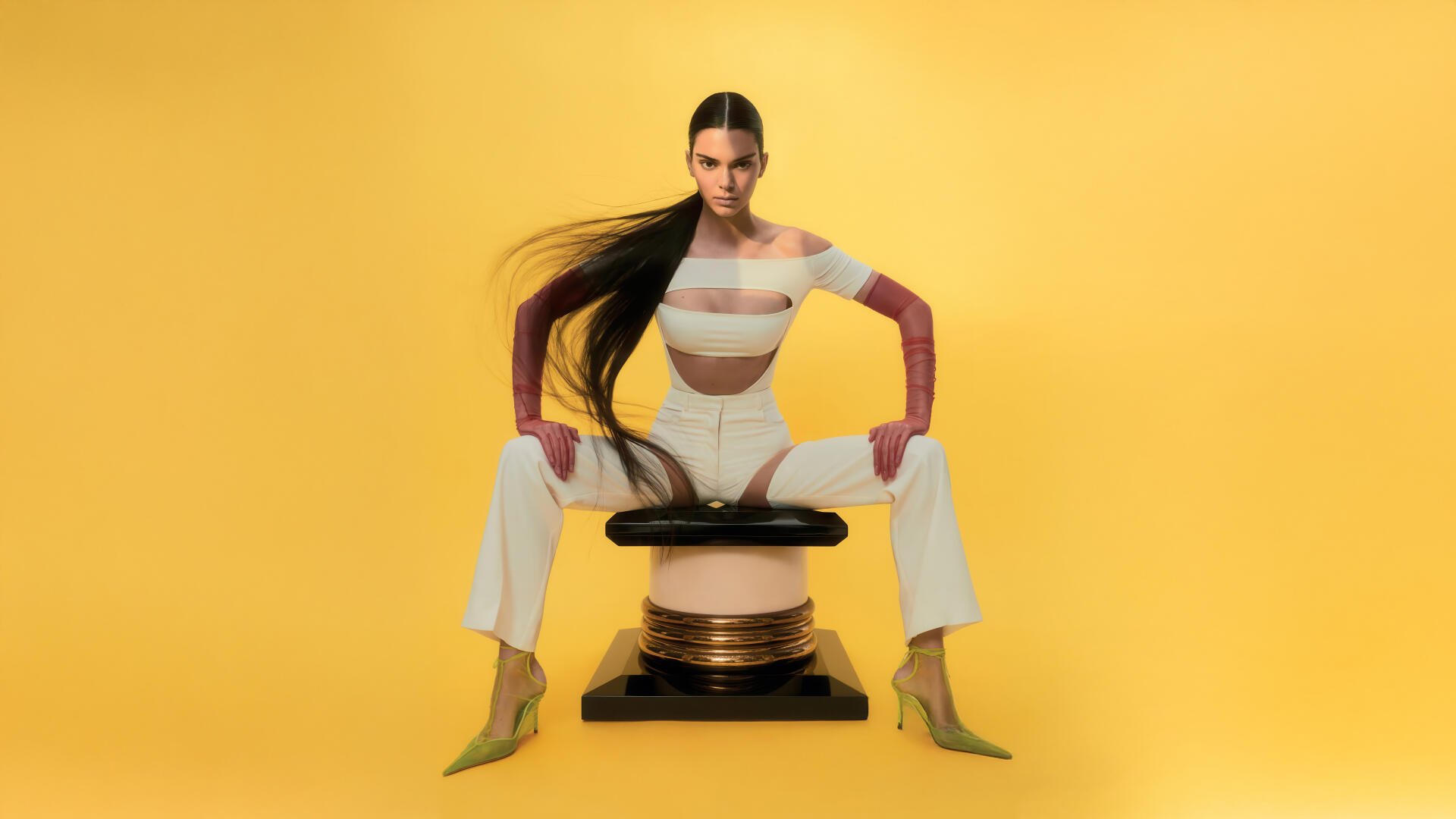 8K Ultra HD PC desktop wallpaper of a celebrity model: woman with long dark hair in a white cutout outfit and yellow heels, seated on a sculptural stool against a vivid yellow background.