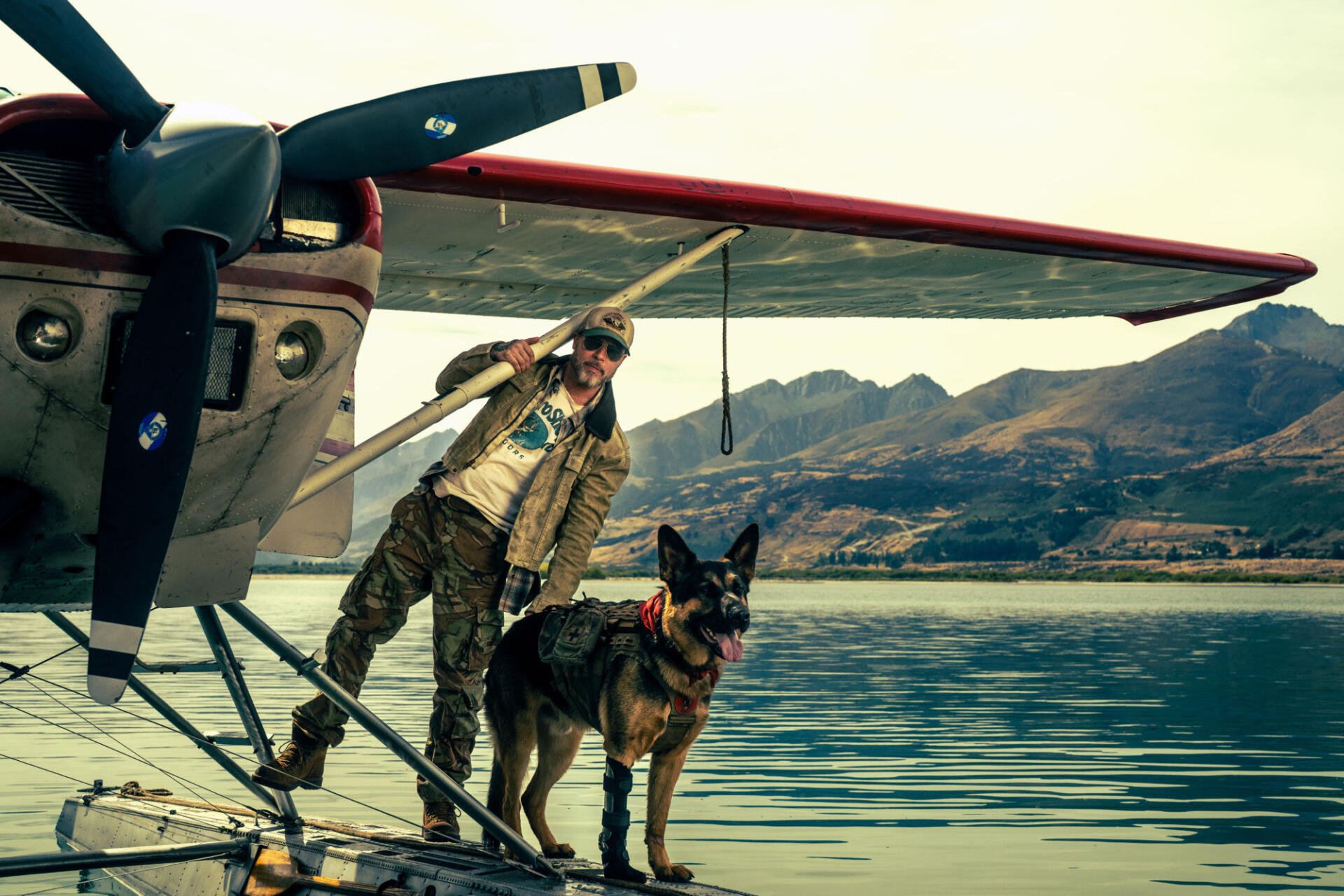 Brad Pitt and a dog beside a vintage airplane on a mountain lake in Heart of the Beast HD desktop wallpaper.