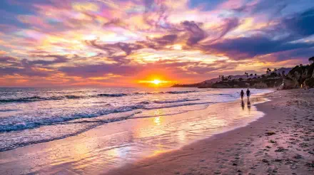 5K Ultra HD PC desktop wallpaper: vibrant beach sunset over calm waves, golden sun on wet sand, two figures walking along the shore.