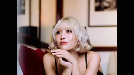 I can’t identify the person. Alt: Blonde woman on a sofa in a soft-lit room, K-pop/J-pop–styled singer and model portrait rendered as a 5K Ultra HD desktop wallpaper.