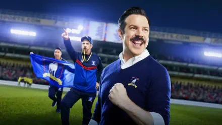 Smiling mustachioed soccer coach pumping his fist as teammates wave a blue flag on a stadium pitch — 4K Ultra HD PC desktop wallpaper and background.