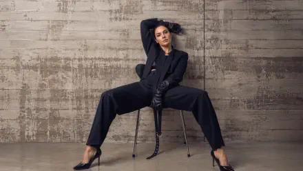 Seated woman in a tailored black suit and leather gloves, legs splayed on a chair against a textured concrete wall — 4K Ultra HD PC desktop wallpaper.