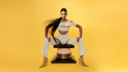 8K Ultra HD PC desktop wallpaper of a celebrity model: woman with long dark hair in a white cutout outfit and yellow heels, seated on a sculptural stool against a vivid yellow background.