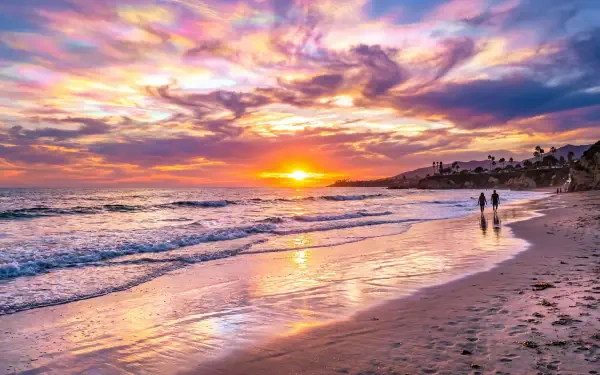 5K Ultra HD PC desktop wallpaper: vibrant beach sunset over calm waves, golden sun on wet sand, two figures walking along the shore.