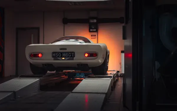 Rear view of a white McLaren G6GT vehicle on a garage lift, warm backlighting and reflections — 8K Ultra HD PC desktop wallpaper and background.