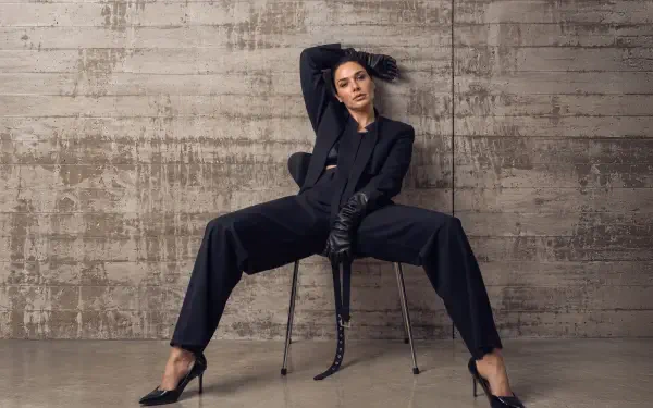 Seated woman in a tailored black suit and leather gloves, legs splayed on a chair against a textured concrete wall — 4K Ultra HD PC desktop wallpaper.