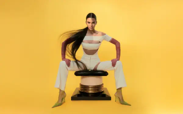 8K Ultra HD PC desktop wallpaper of a celebrity model: woman with long dark hair in a white cutout outfit and yellow heels, seated on a sculptural stool against a vivid yellow background.