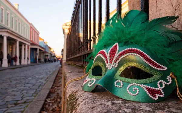 Green feathered Mardi Gras carnival mask on a stone ledge with a blurred New Orleans cobblestone street, 2K Quad HD PC desktop wallpaper and background.